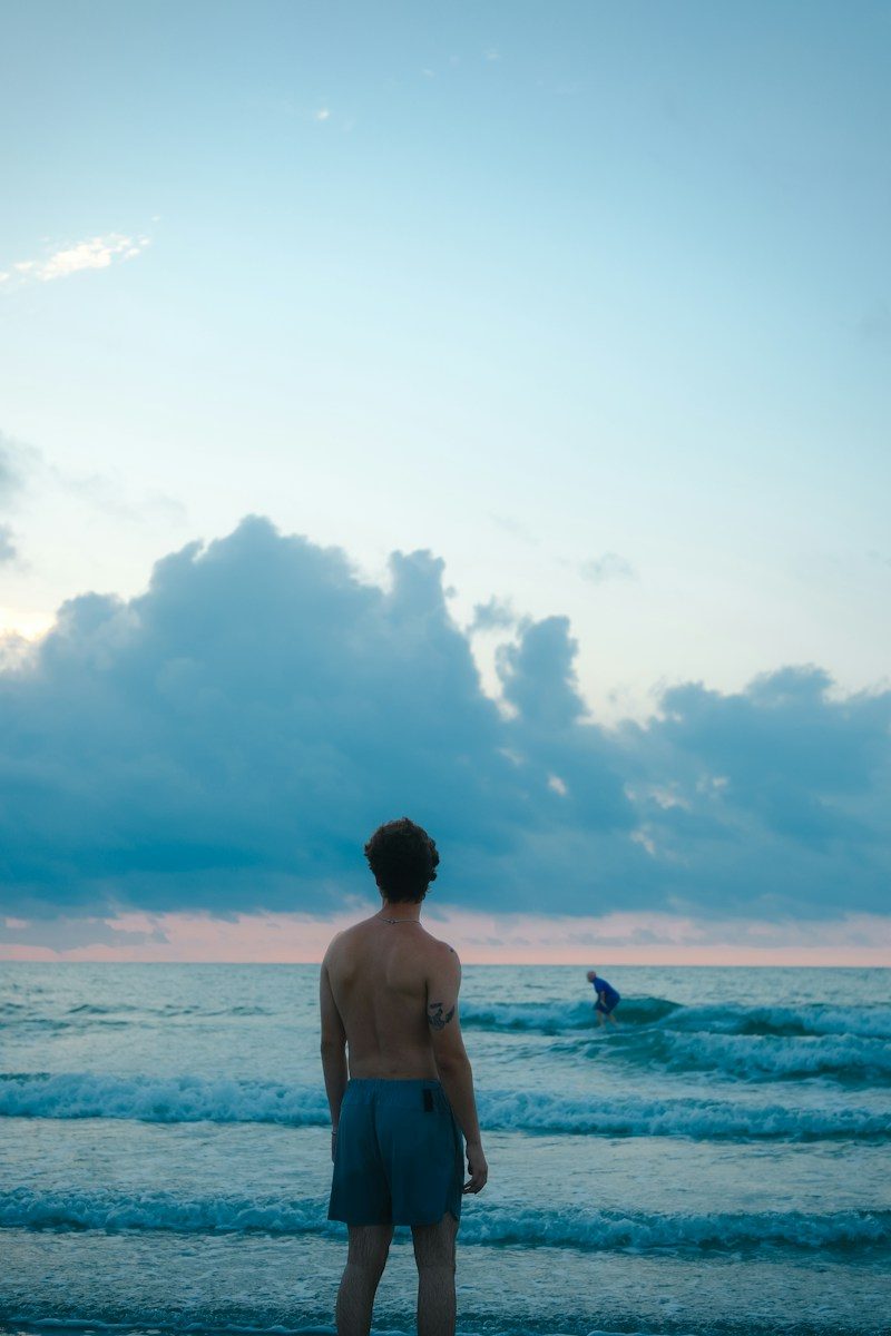 A shirtless man in blue shorts looking at the ocean under a cloudy sky