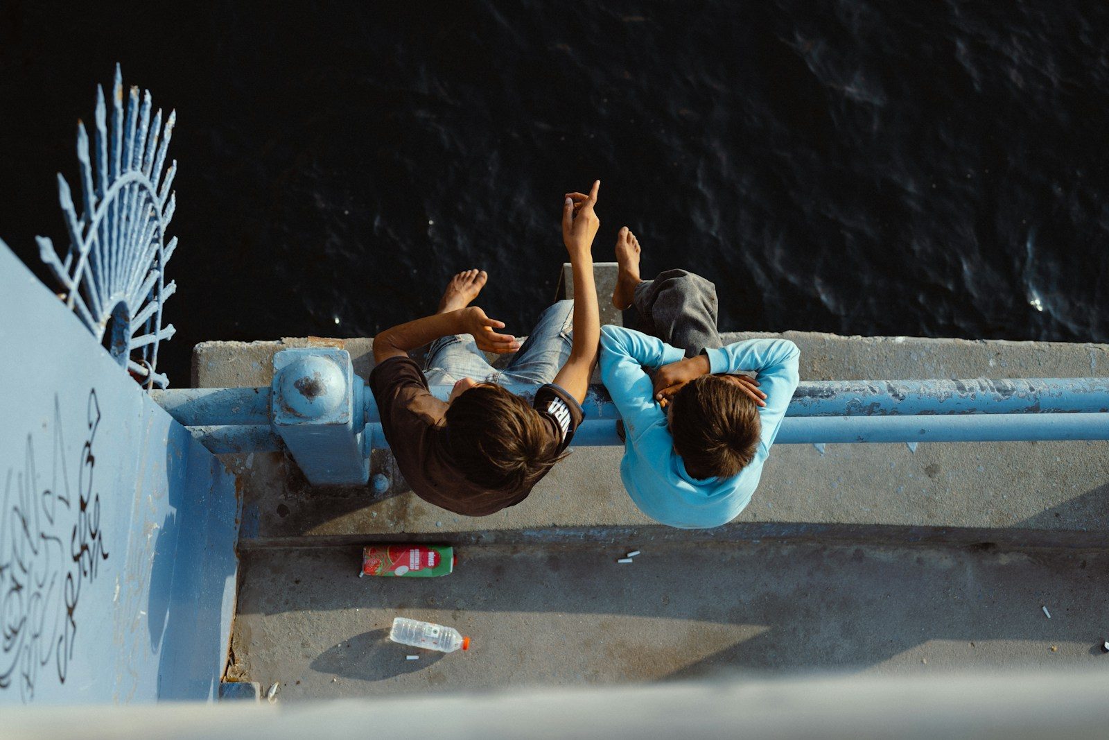Two people sitting on a blue bridge railing pointing toward dark water below